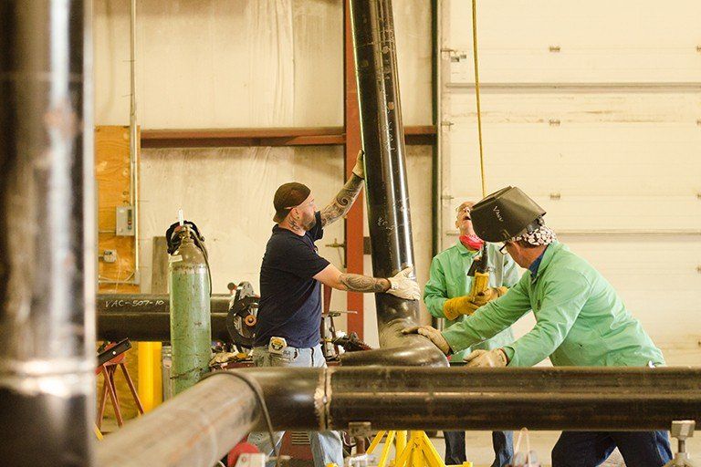 Three people working on black pipes in a workshop. One welds while another assists, one supports the pipe.