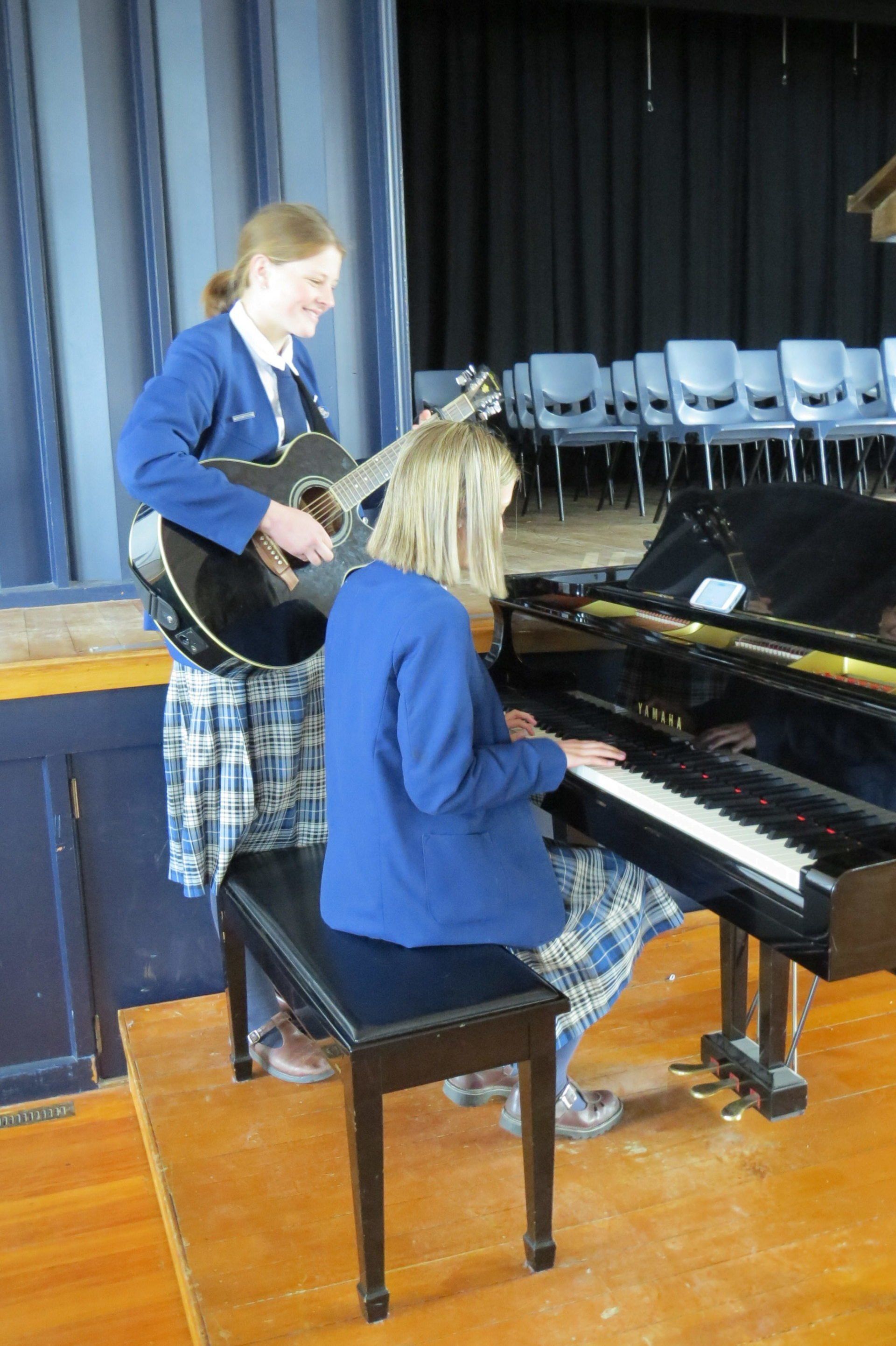 A girl is playing a guitar and another girl is playing a piano.