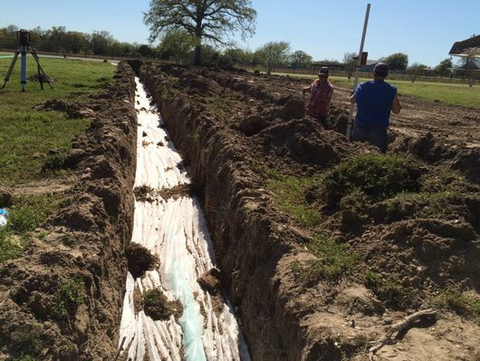 Digging a long irrigation trench in a grassy field, with workers standing beside it under a clear sky.