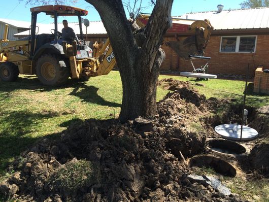 Backhoe at a yard digging around a tree beside a brick house and grassy yard