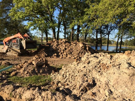 Excavator beside large dirt piles near trees and a lake on a sunny day
