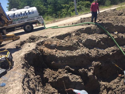 Collapsed dirt trench at a construction site with a worker standing nearby and a green hose across the ground