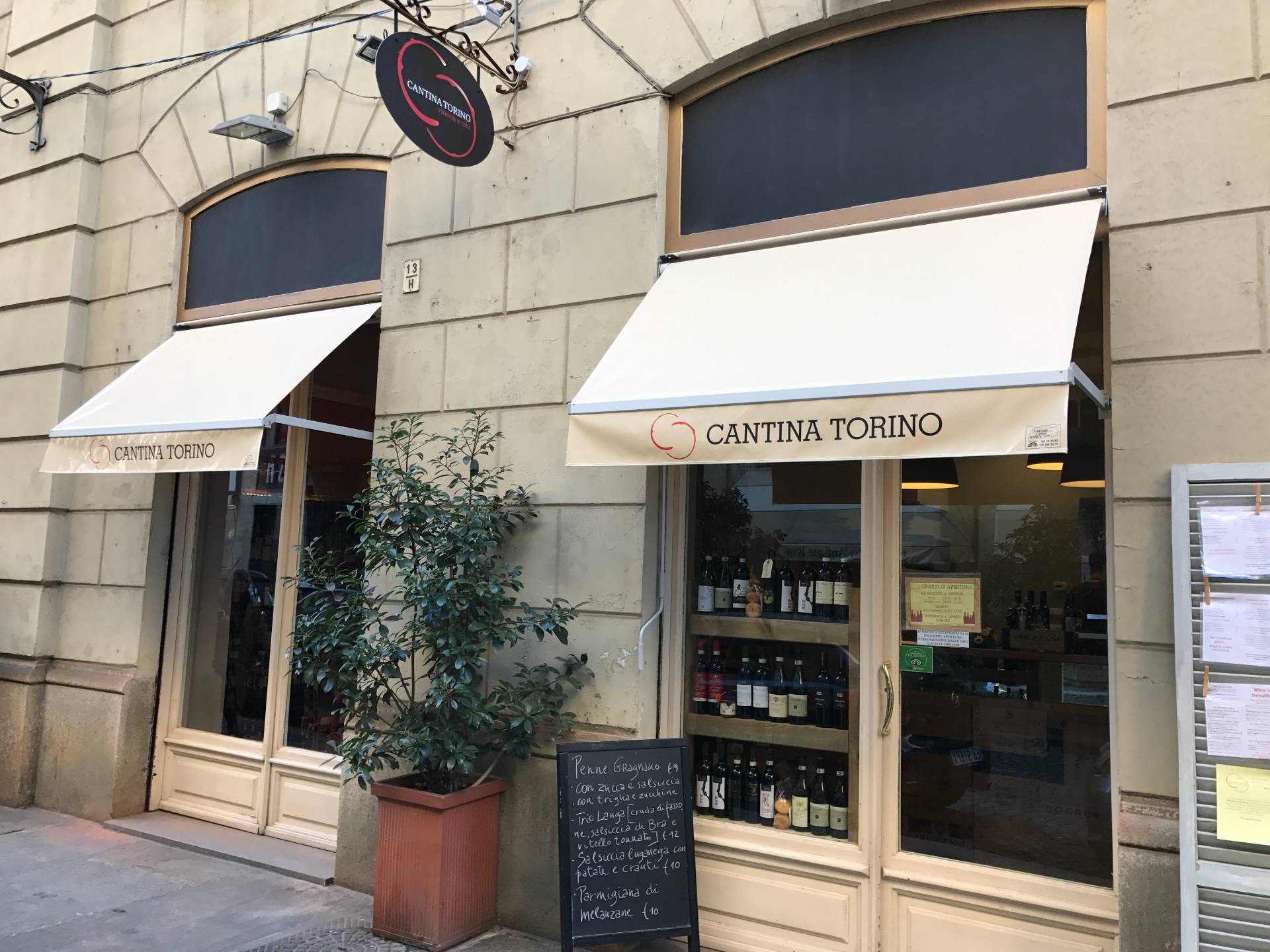 Vetrina della Cantina Torino con tende alla pantalera, porta in vetro, bottiglie di vino a vista e menù scritto sulla lavagna.