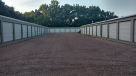 A view down a gravel alleyway lined with white self-storage units under a line of trees.