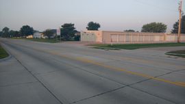 A paved road with yellow center lines leading to a small, tan self-storage facility under a clear, pale sky.