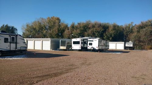 Several white recreational vehicles parked in a gravel lot next to a line of storage units with trees in the background.