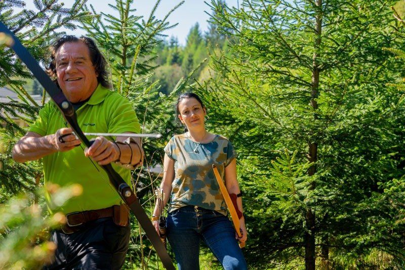 Ein Mann zielt mit Pfeil und Bogen, eine Frau steht daneben im Wald; beide lächeln.