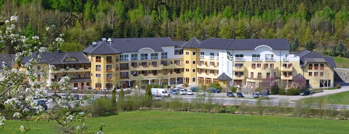 Großes gelb-weißes Gebäude mit dunklen Dächern vor einem grünen Feld, umgeben von Bäumen.
