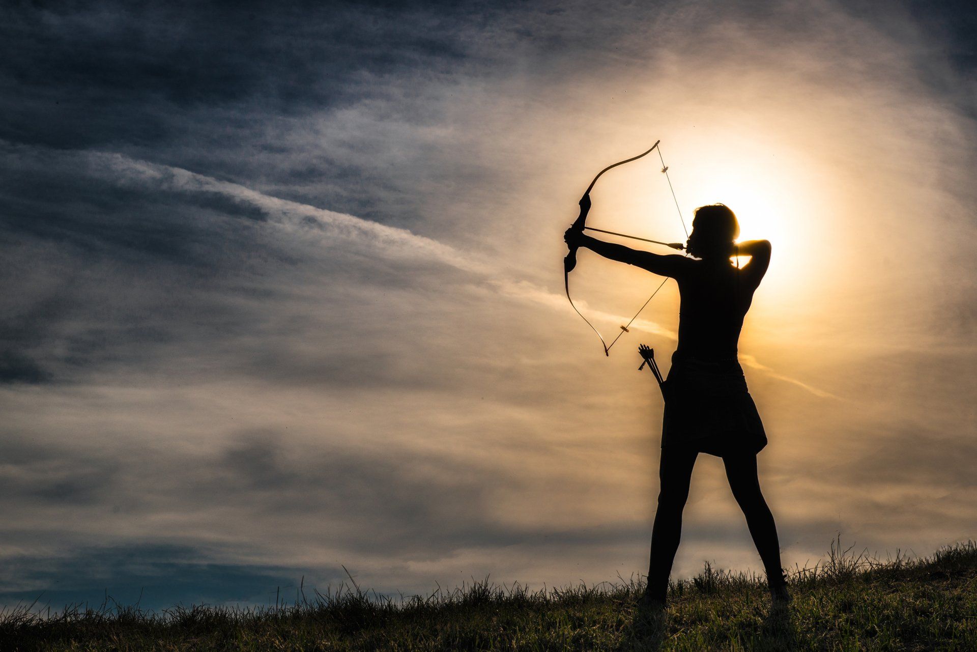 Silhouette einer Person, die Pfeil und Bogen spannt, von der Sonne hinterleuchtet vor einem bewölkten Himmel.