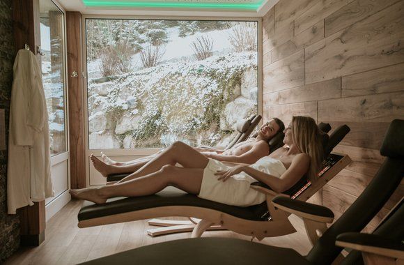 Zwei Personen entspannen sich auf Liegestühlen in einem Spa und blicken aus dem Fenster auf den Schnee.