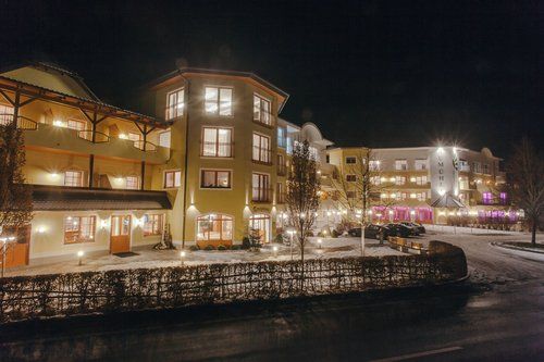 Hotelaußenansicht bei Nacht, beleuchtete Fenster, beiges und gelbes Gebäude, schneebedeckter Boden, kleine Lichter entlang des Weges.