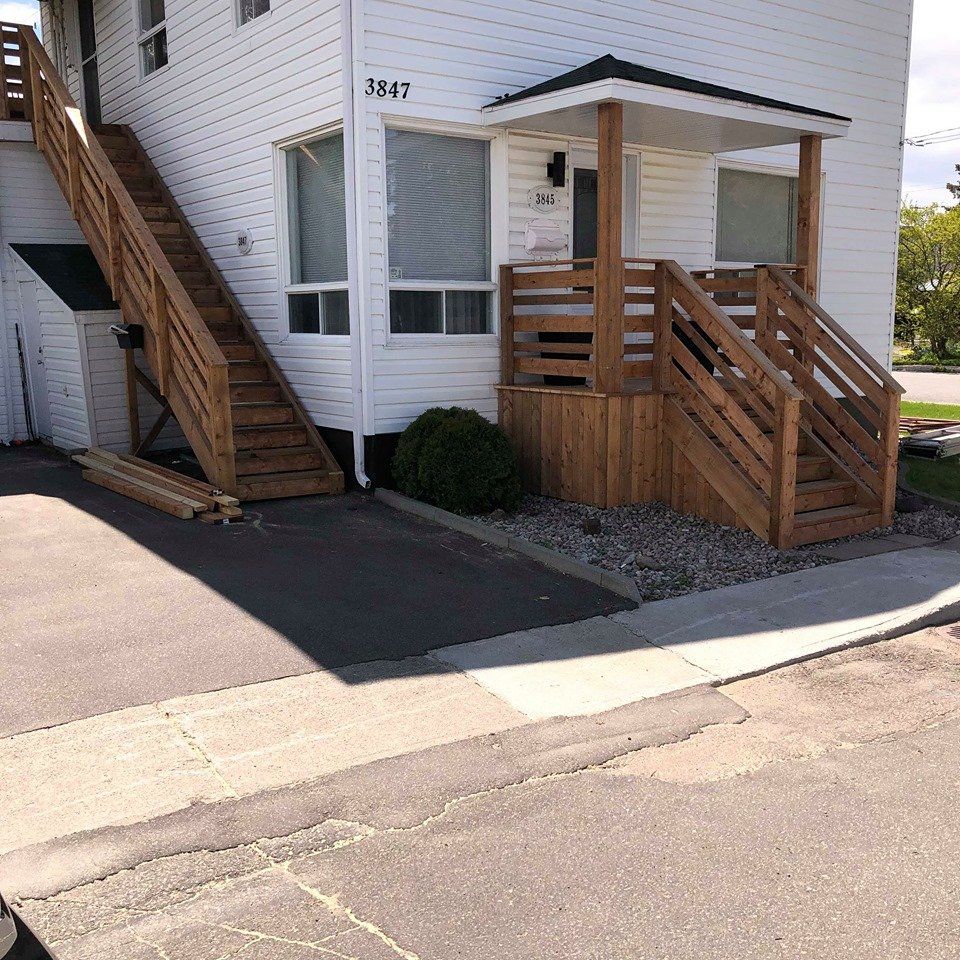 Bâtiment blanc de deux étages avec escalier et rampe en bois. Une allée et un trottoir en asphalte noir se trouvent au premier plan.