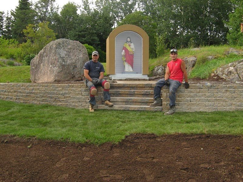 Deux hommes sont assis sur des marches en pierre près d'une fresque religieuse. De l'herbe verte et des arbres se dessinent à l'arrière-plan.