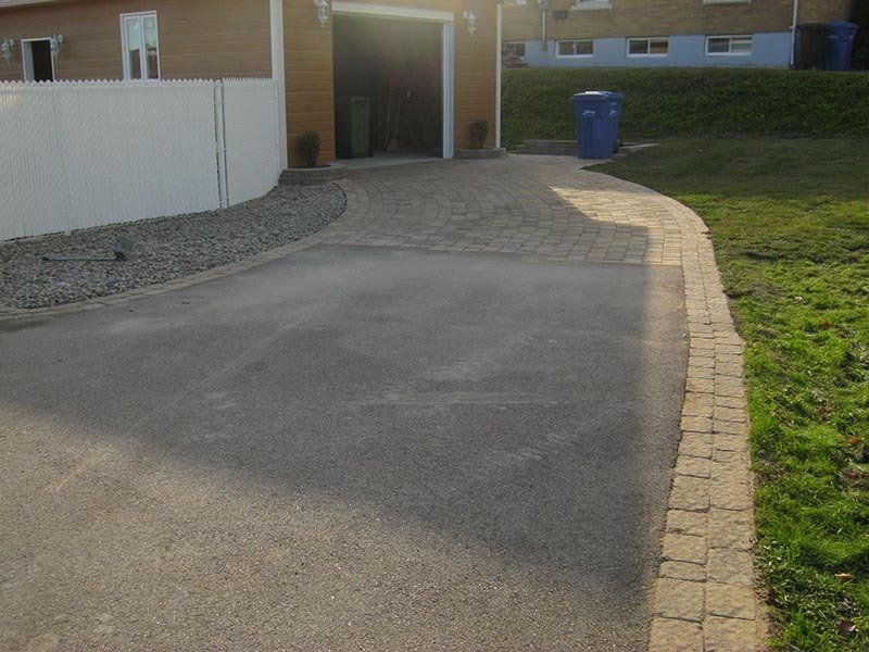 Allée asphaltée bordée de pierres et de gravier menant à un garage ; pelouse à droite.