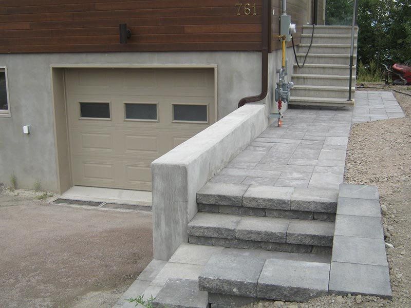 Vue extérieure d'une maison avec garage, escalier et allée pavée. Éléments en béton et en pierre.
