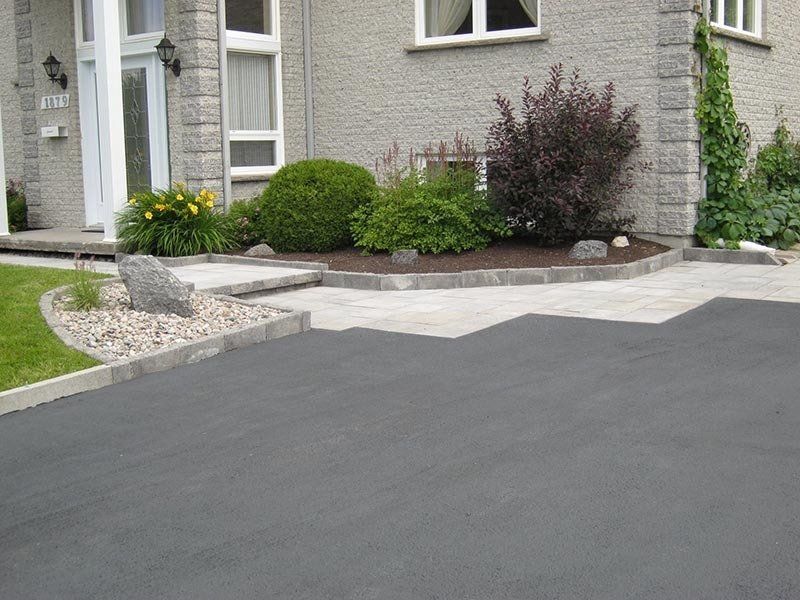 Allée en asphalte gris menant à une maison avec un parterre de jardin aménagé.