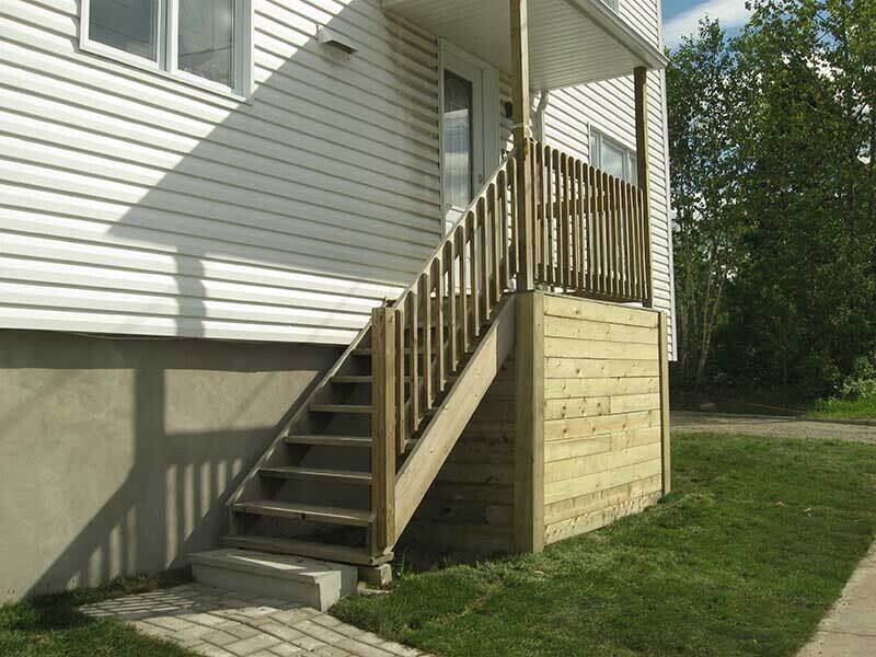 Un escalier extérieur en bois mène à un porche et à la porte d'un bâtiment aux murs blancs, bordé d'herbe verte.