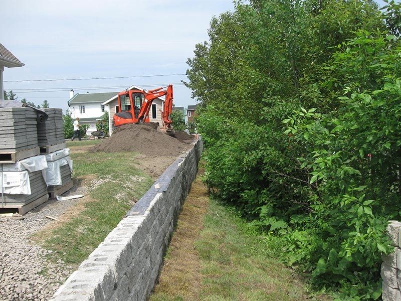 Chantier : pelleteuse orange sur la terre, mur de parpaings gris, buissons verts et maisons en arrière-plan.