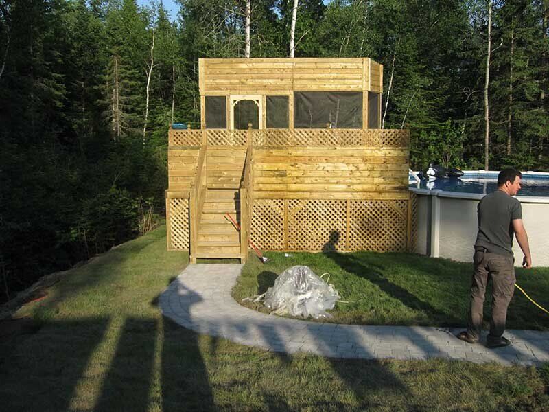 Terrasse en bois à plusieurs niveaux avec escalier, treillis et toit grillagé. Un homme avec un tuyau d'arrosage près d'une piscine. Arbres en arrière-plan.