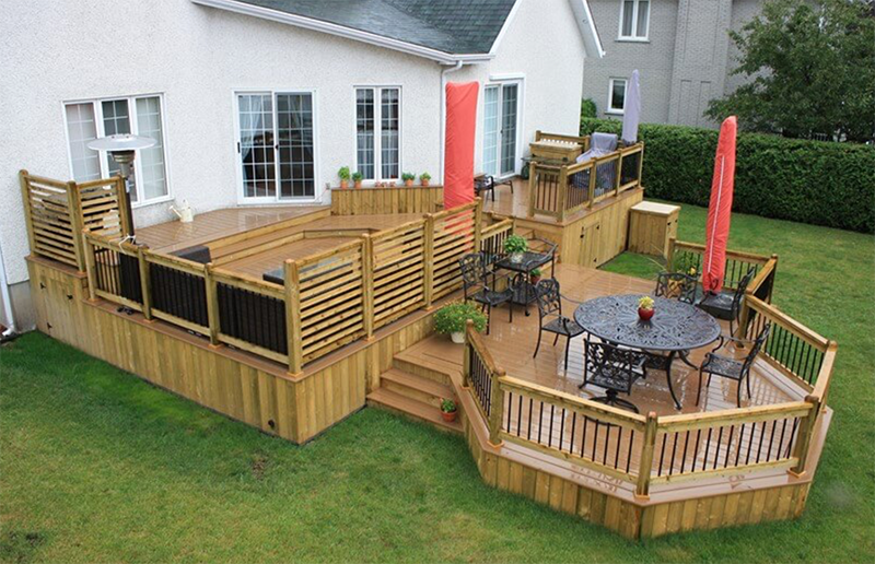 Terrasse en bois à plusieurs niveaux attenante à une maison blanche, avec rambardes, mobilier et parasol rouge.