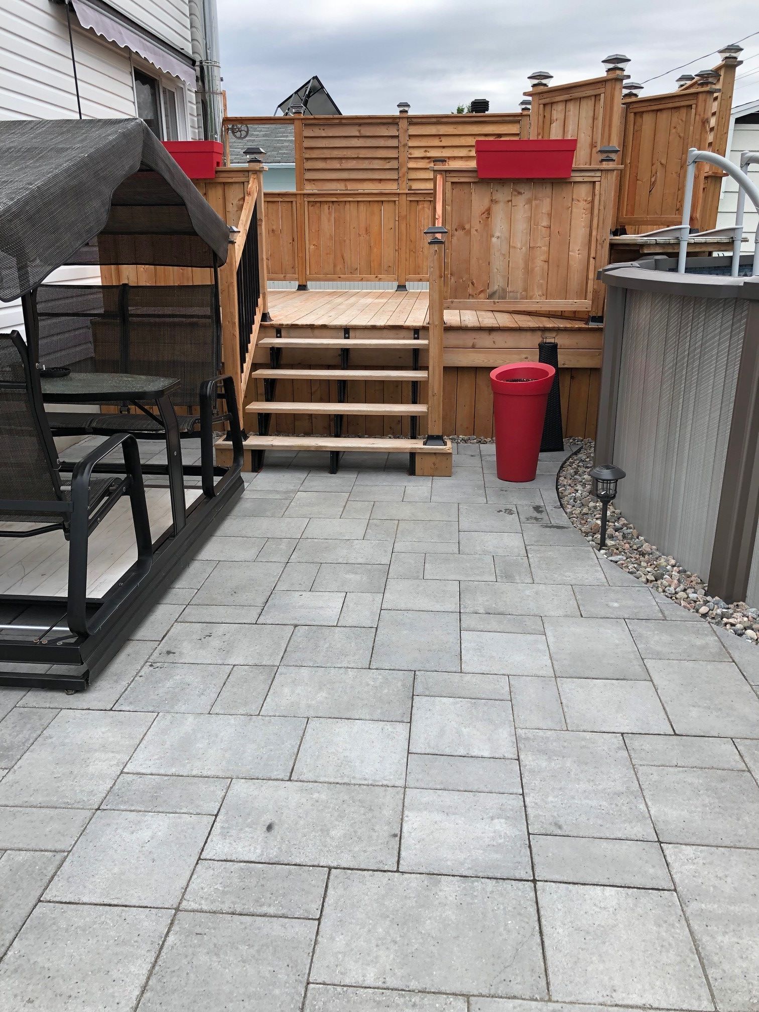 Terrasse pavée de dalles grises, marches menant à une terrasse en bois avec piscine, gazebo sur la gauche.
