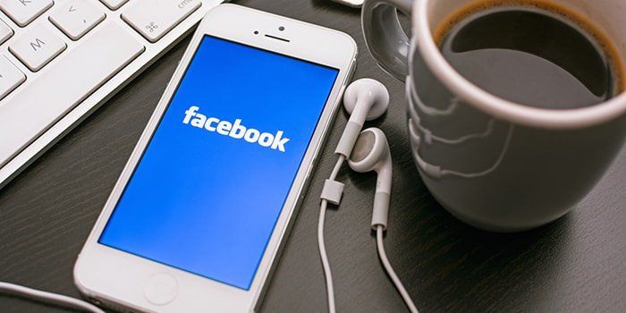 A cell phone with facebook on the screen next to a cup of coffee