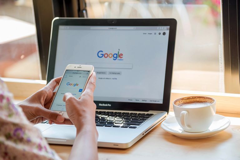 A person is using a cell phone in front of a laptop that says google on the screen