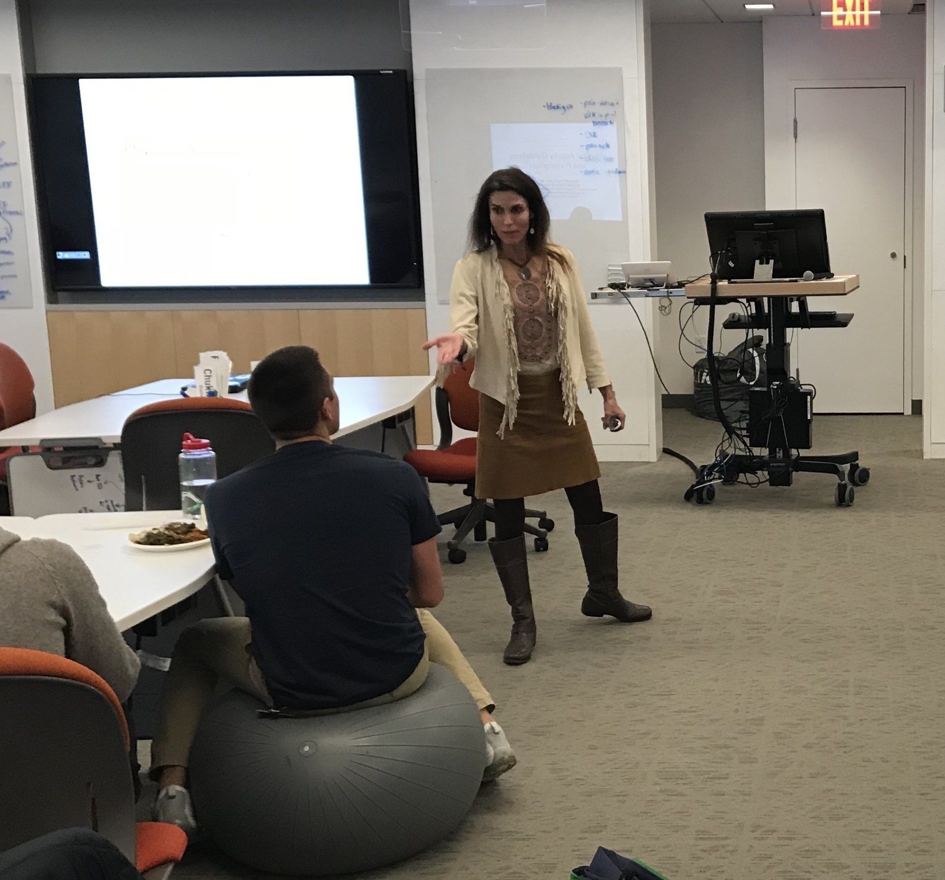Woman gesturing, speaking in a classroom. A man sits on an exercise ball, facing away. Whiteboard and monitor in background.