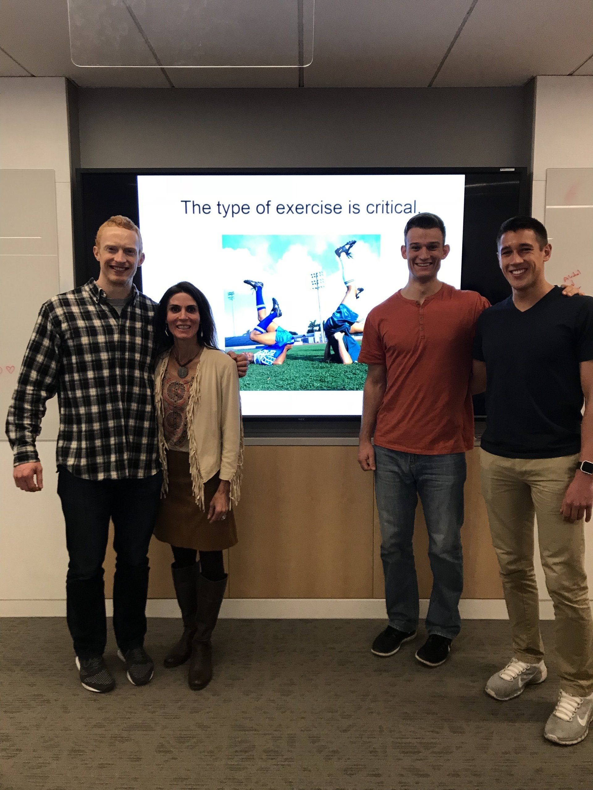 Four people stand in front of a screen displaying a presentation. The screen shows a picture of people playing outside.