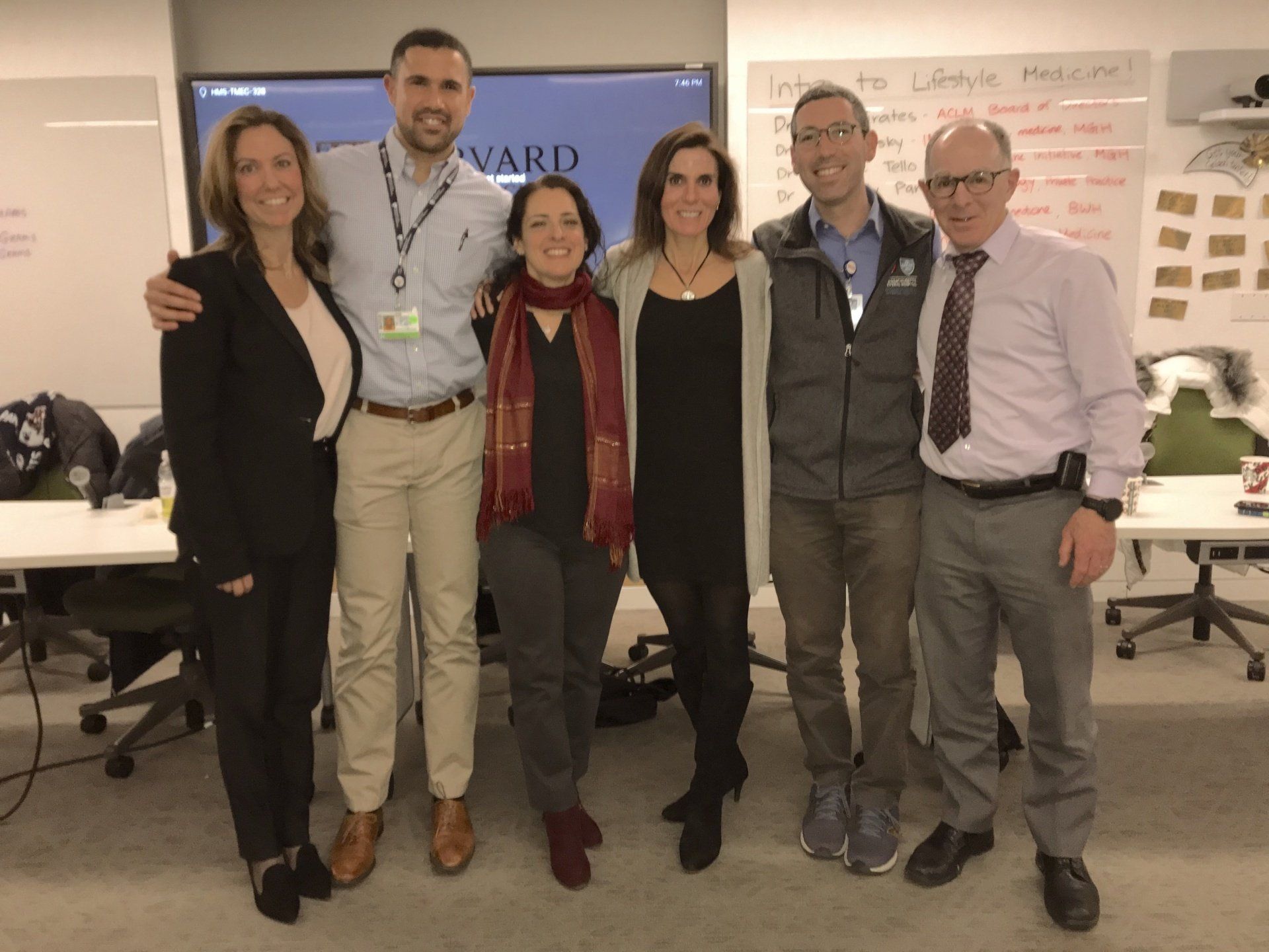 Six people smiling, posing together indoors near a screen and sticky notes.