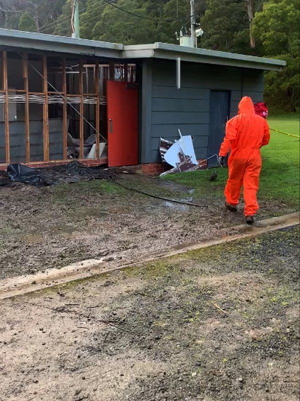 A person in an orange hazmat suit walks near a partially demolished gray building with a red door.