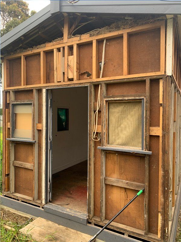 Exterior of a shed under renovation. The wooden frame is exposed, and doorway leads into a white-walled room with a painting.
