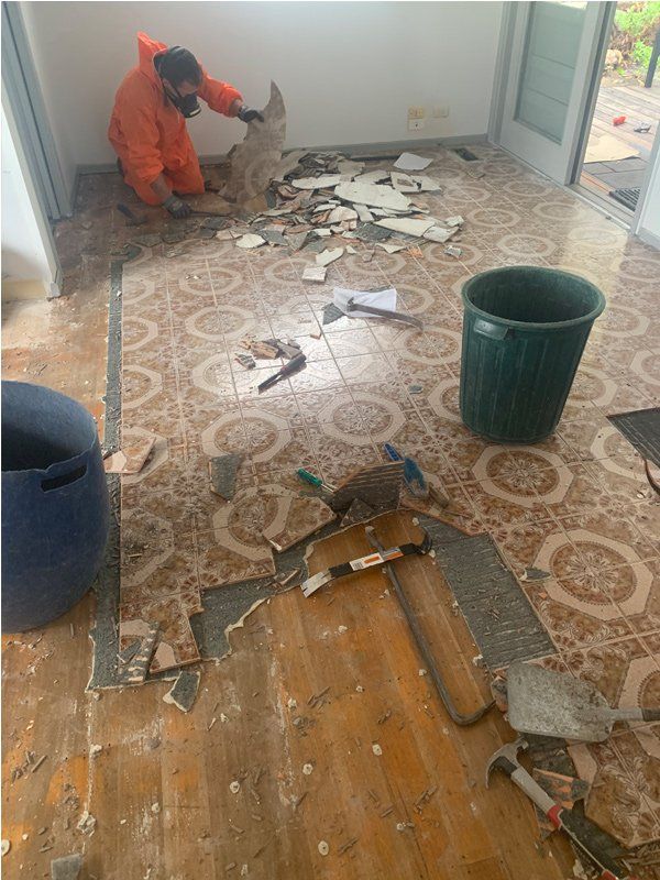 Person in orange suit removing tile floor in a room. Debris and tools are scattered around.