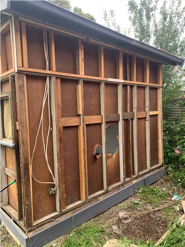 Exterior wall of a shed with exposed wood framing and weathered siding. Wires hang down near an electrical outlet.