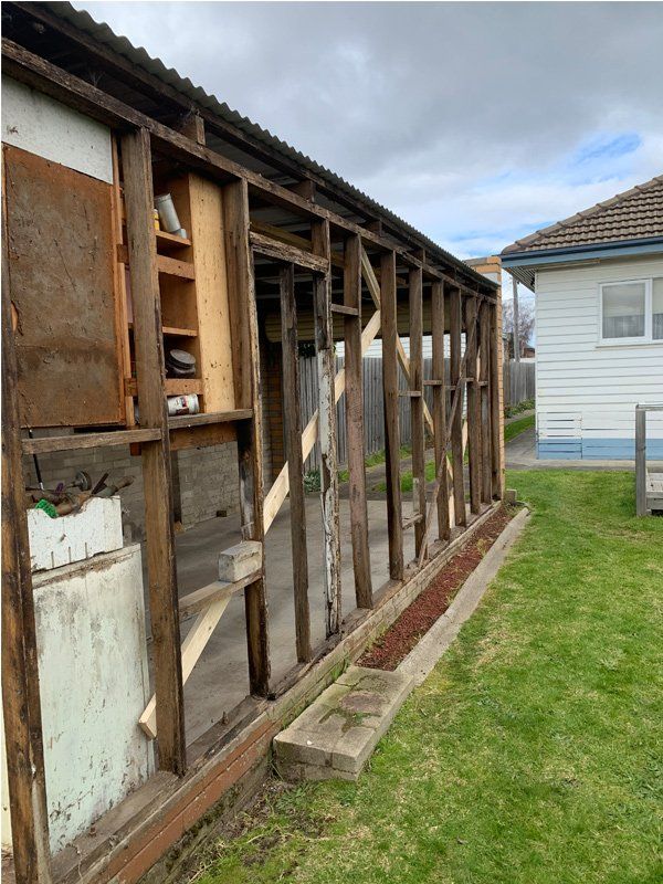 Exterior view of a dilapidated wooden building frame. Green grass and a residential home are in the background.
