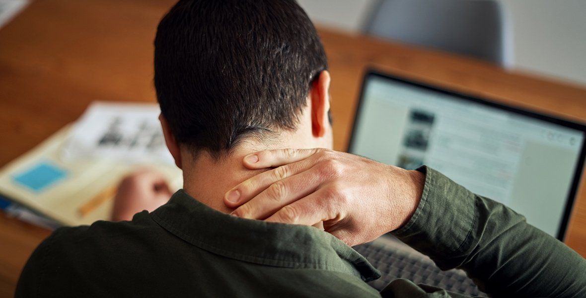 Man seated at a desk, holding his neck, with a laptop and newspaper visible. He appears to be experiencing neck pain.