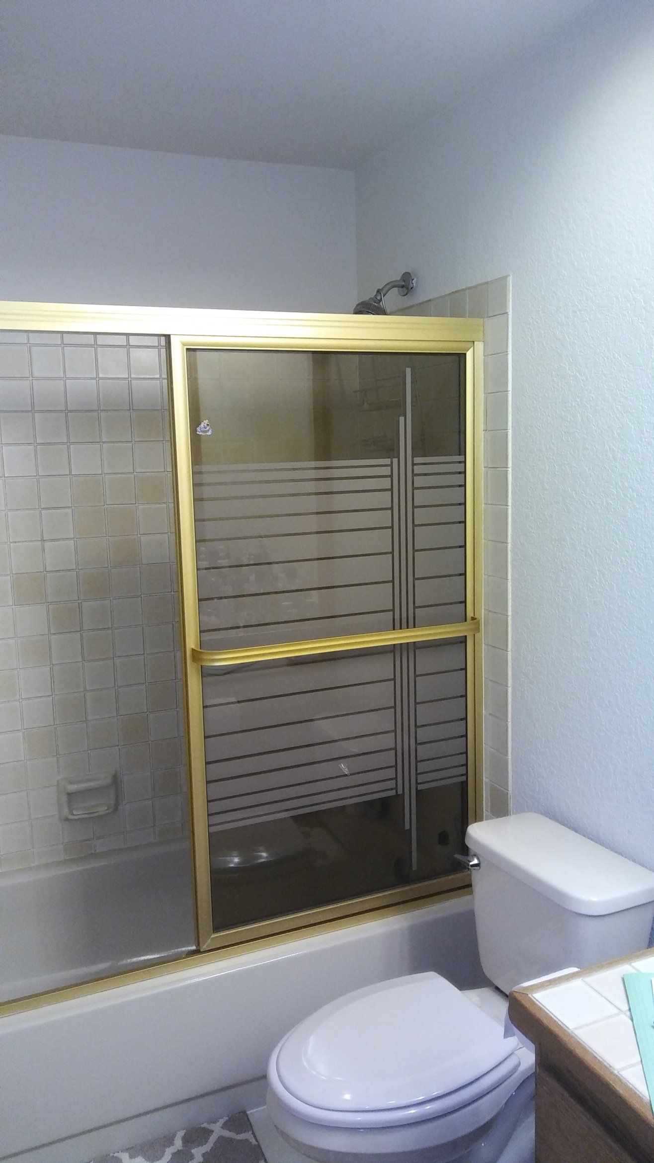 Small bathroom with a gold-framed shower enclosure, toilet, and sink beside a frosted window.