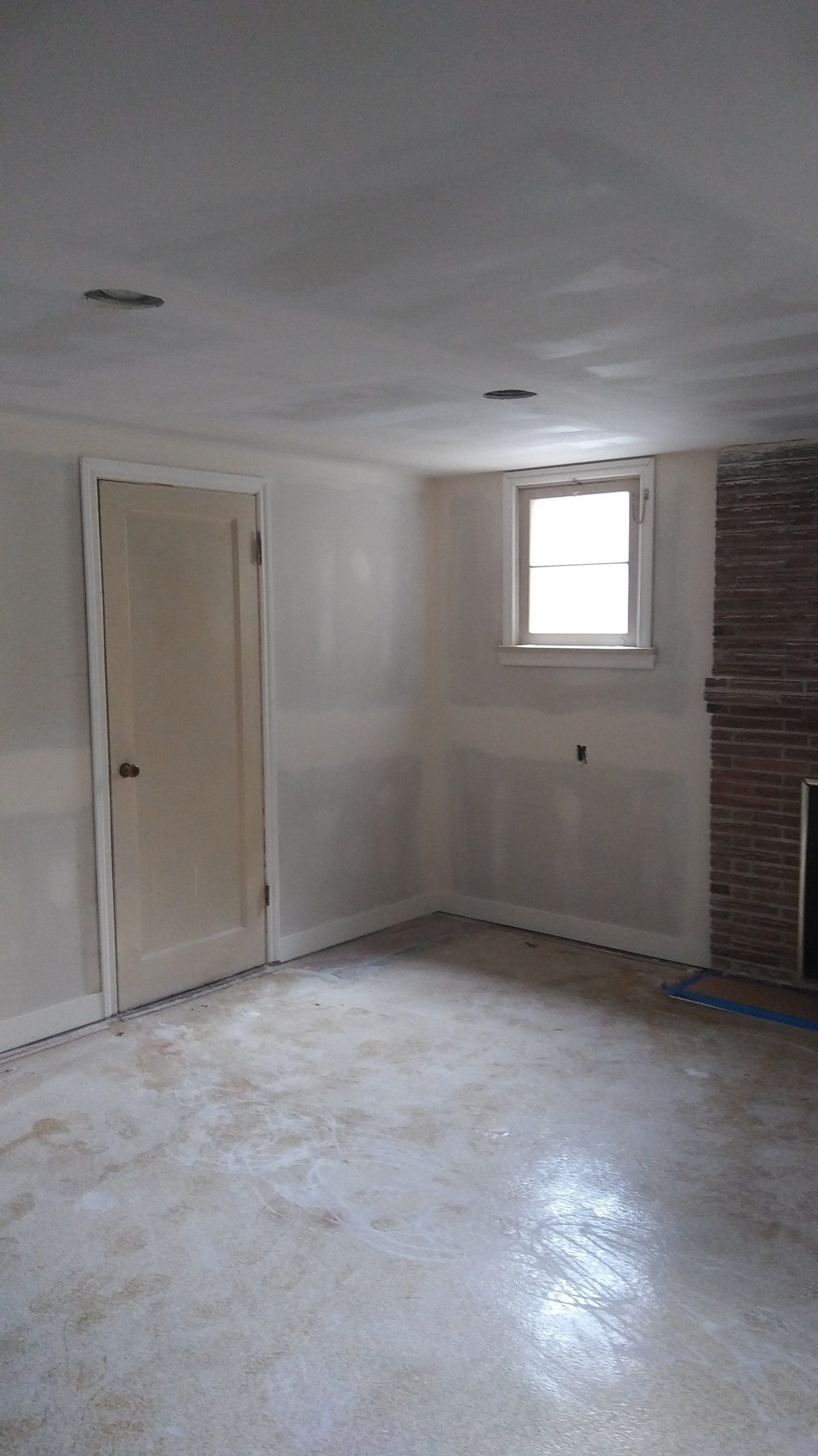 Empty unfinished room with white walls, small window, beige door, and exposed brick wall on the right.