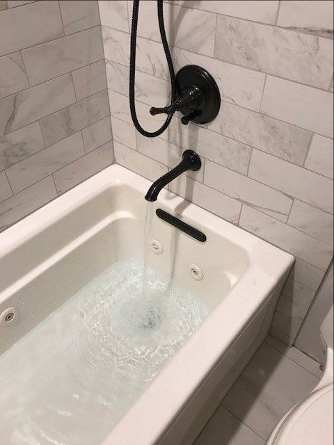 Bathtub with a black shower fixture and handheld sprayer filling with running water in a tiled bathroom