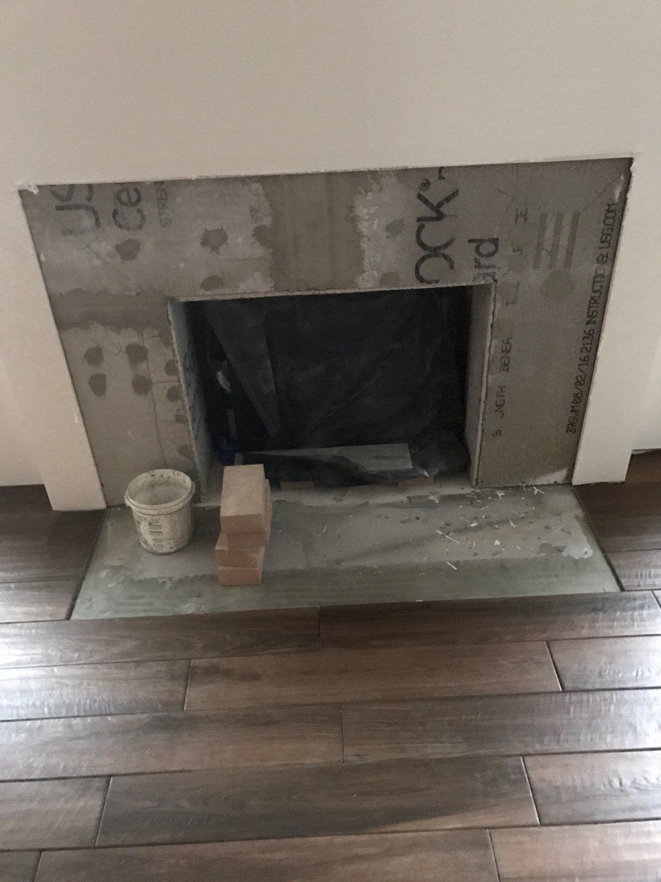 Unfinished fireplace opening with gray cement board, small cup on the left, and dark wood flooring.