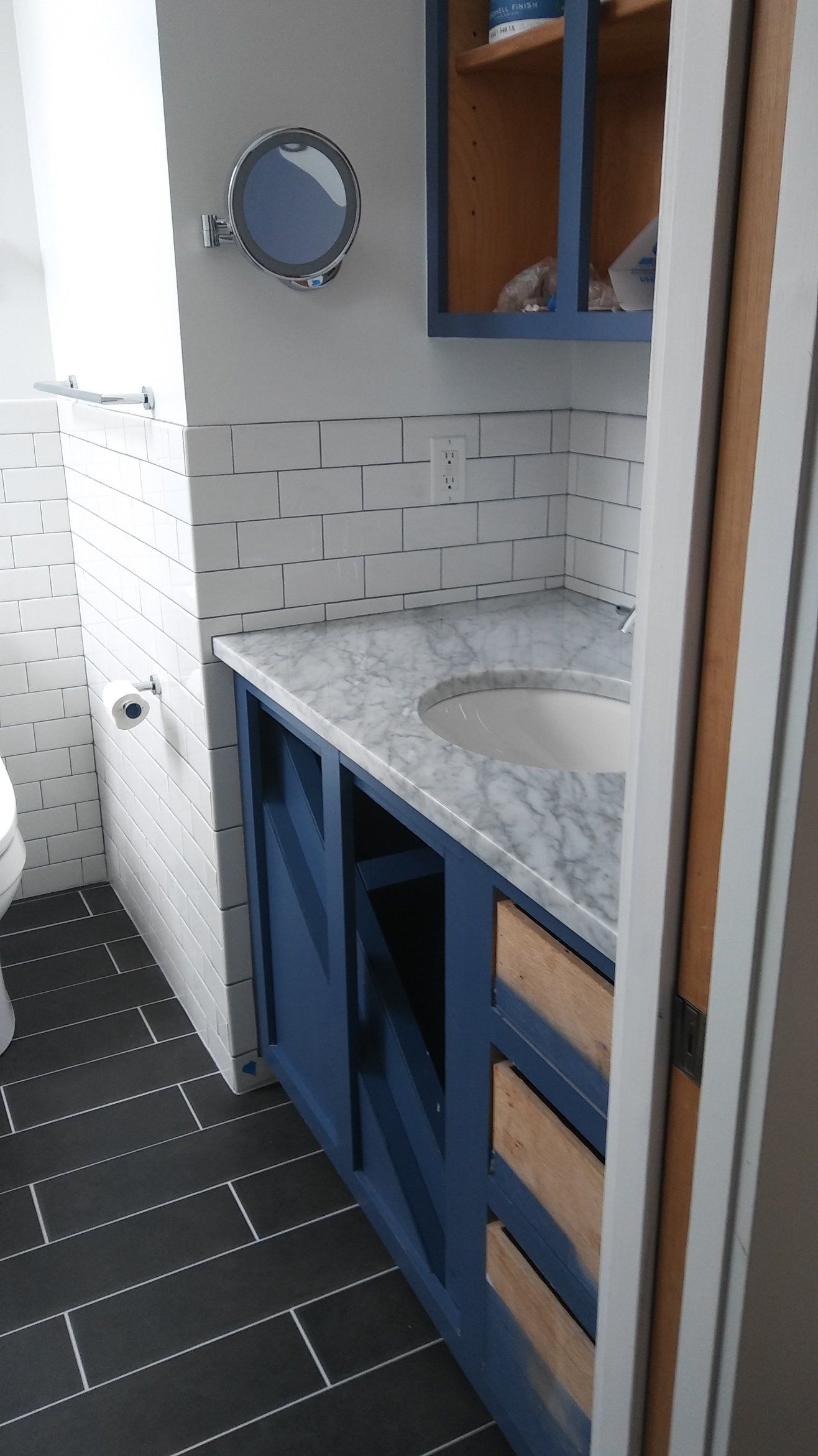 Small bathroom vanity with blue cabinets, speckled countertop, round mirror, and tiled walls.