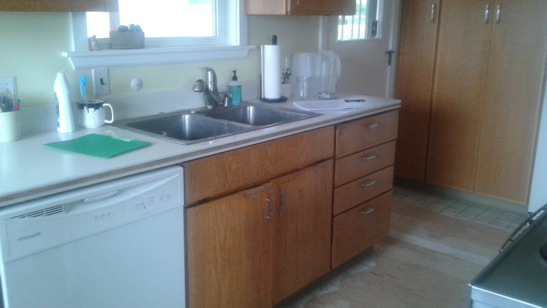 Kitchen with wooden cabinets, sink under window, dishwasher, and countertop items on a tiled floor.