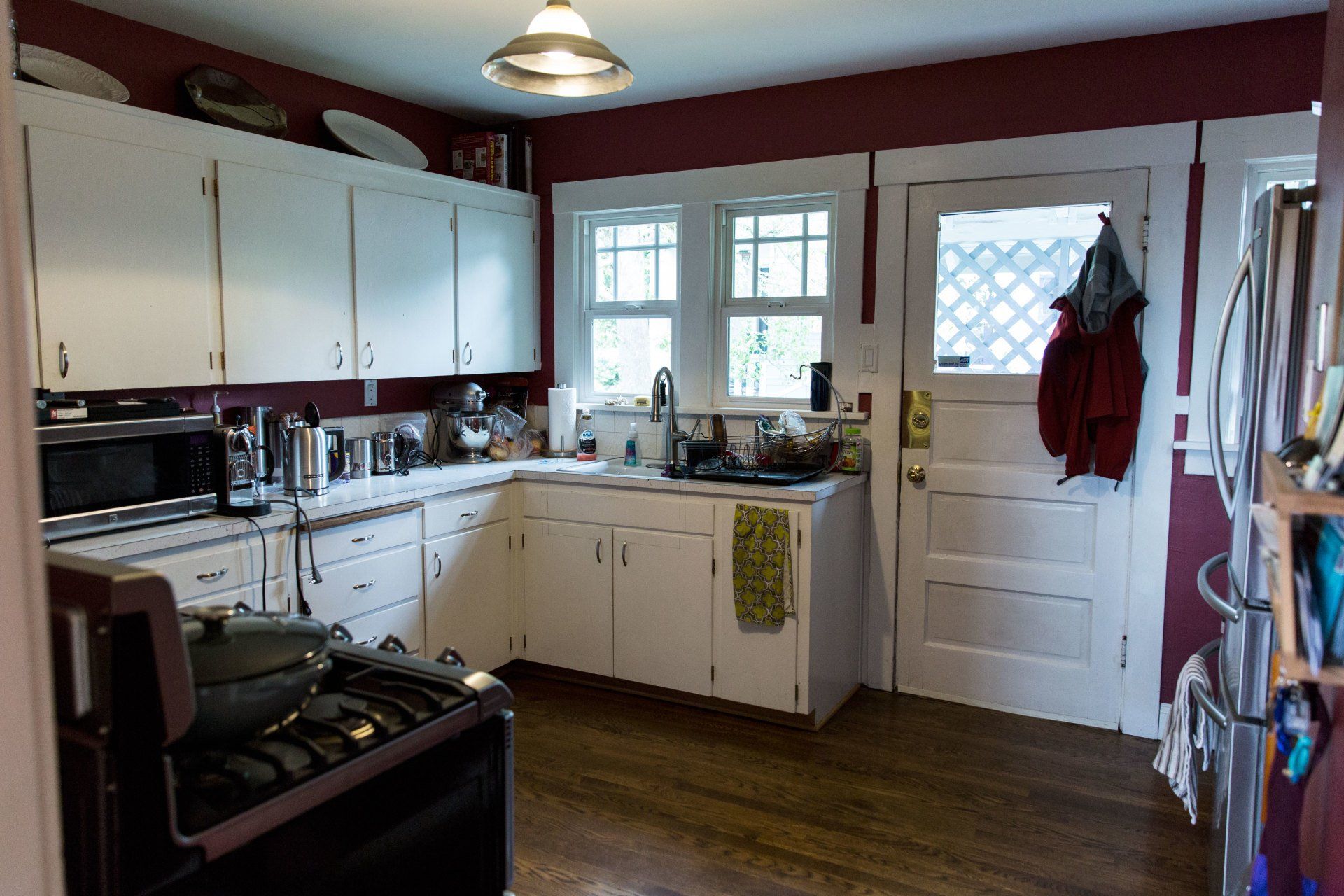 Small kitchen with white cabinets, dark wood floor, a stove, sink, windows, and a door with hanging coats.