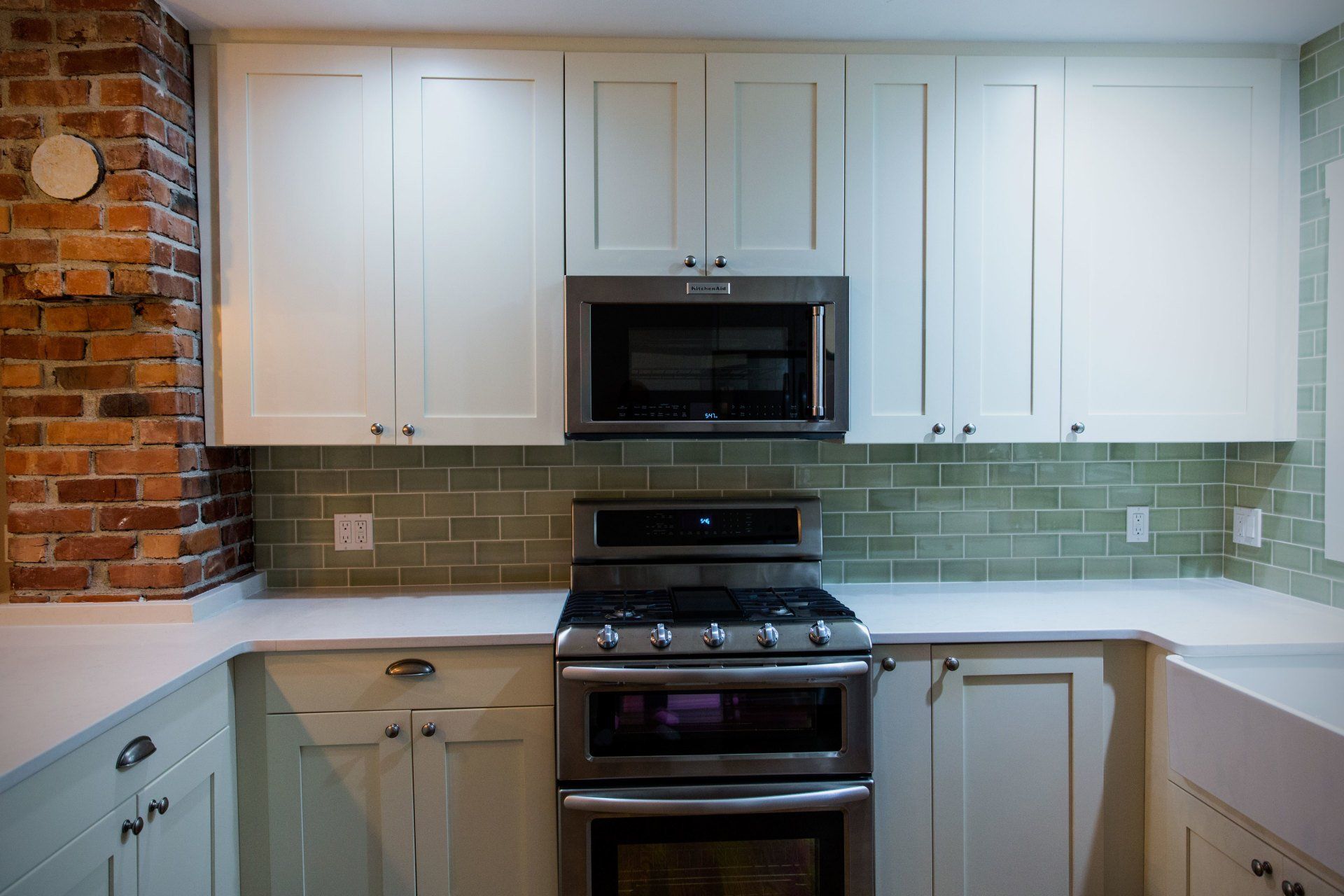 White kitchen with white cabinets, stainless steel stove and microwave, green tile backsplash, and brick wall accent