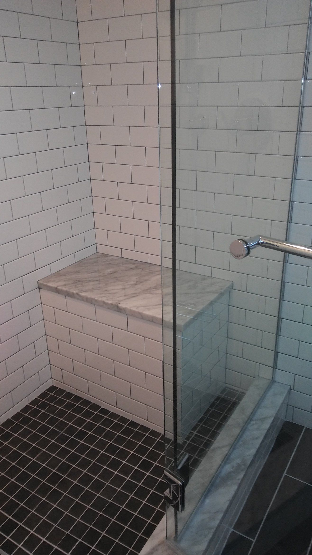 Built-in shower bench with white tile walls and black pebble floor beside glass partition