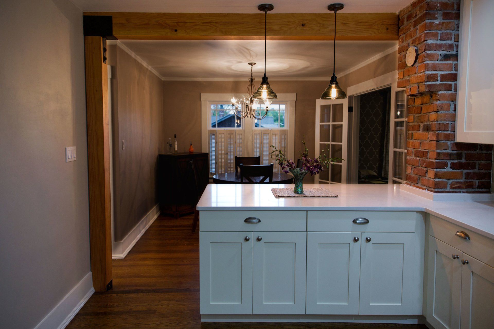 Modern kitchen with white island, pendant lights, hardwood floors, and brick column leading to dining area