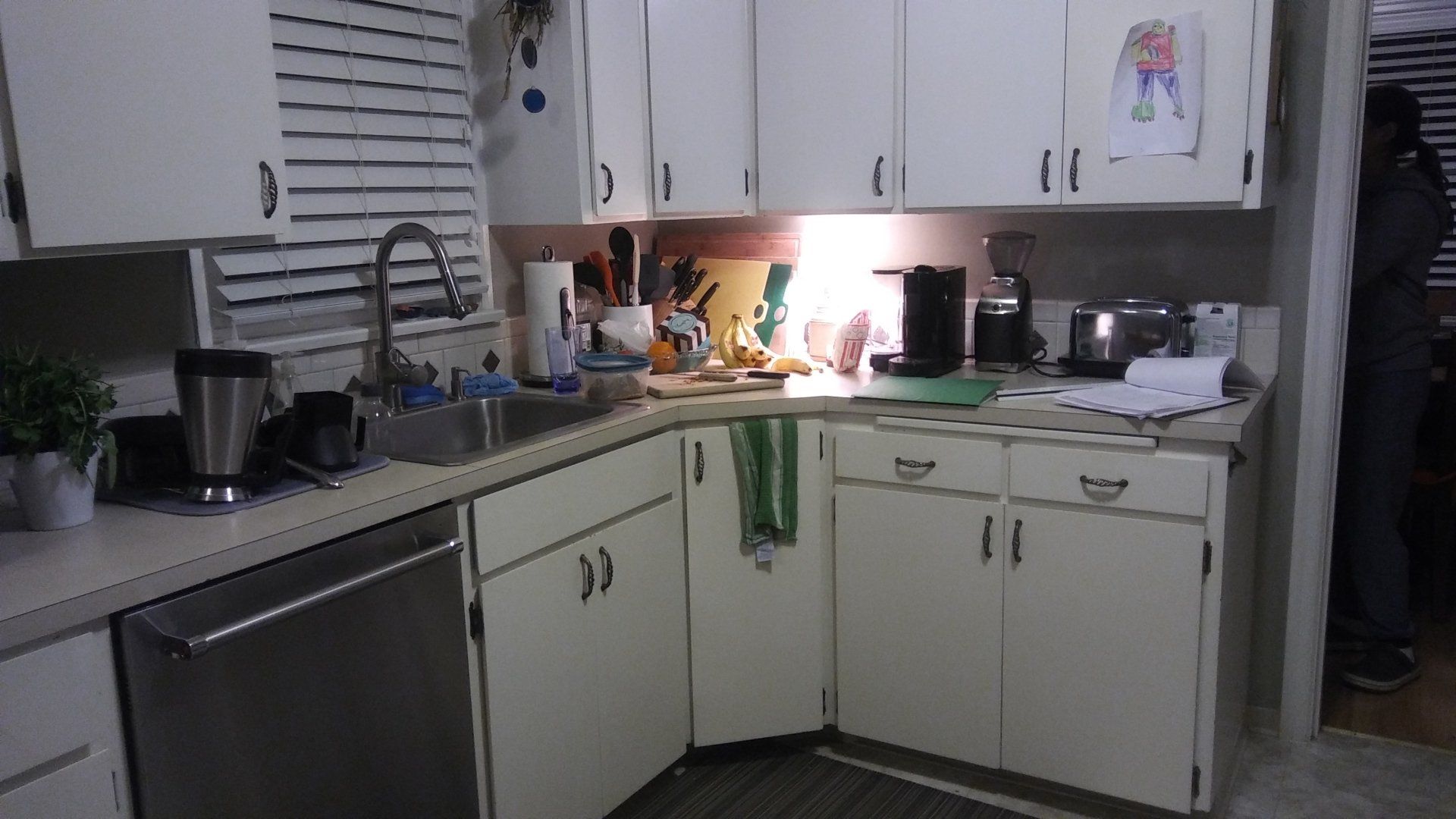 A cluttered kitchen counter with dishes, appliances, and white cabinets under warm lighting.