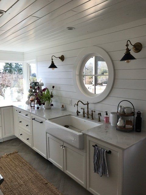 Bright white farmhouse kitchen with apron sink, round window, and dark wall sconces