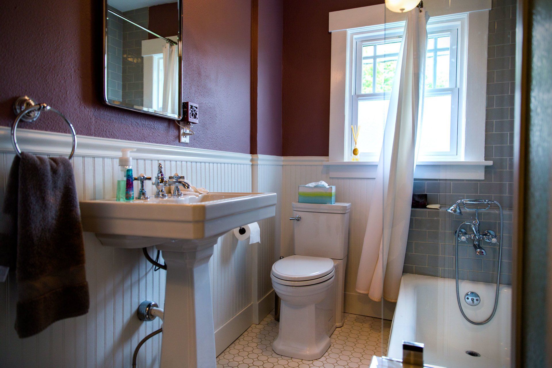 Small bathroom with a pedestal sink, toilet, bathtub, and window with white curtains.