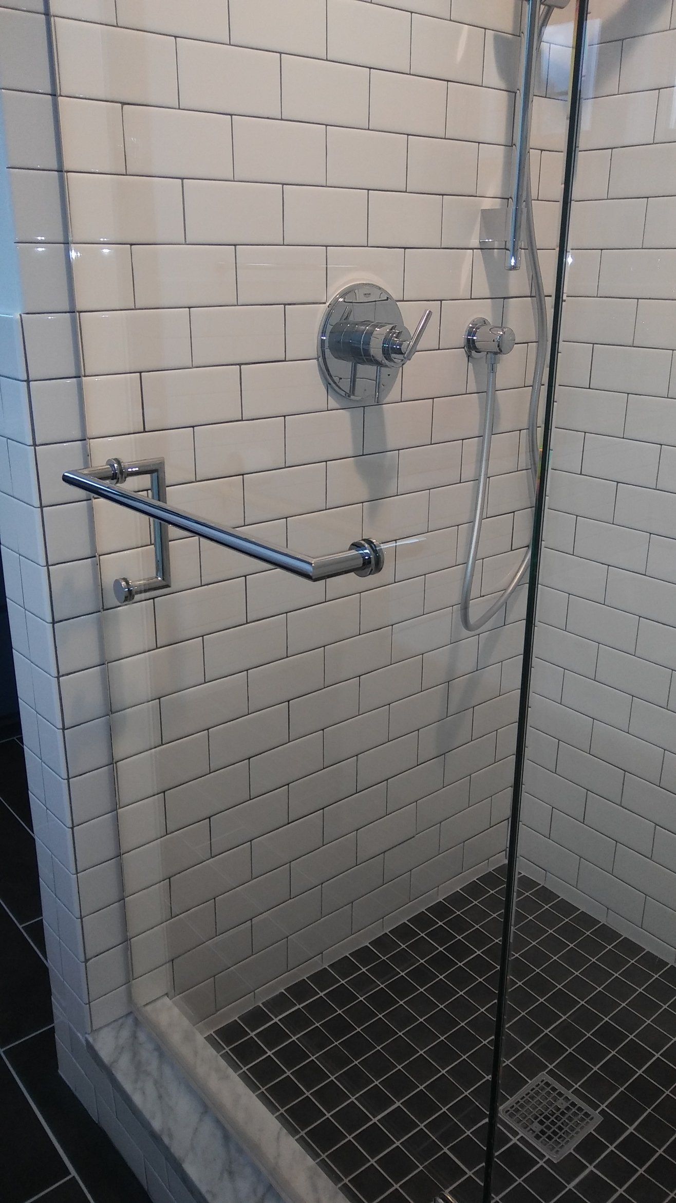 Walk-in shower with glass door, grab bar, rainfall showerhead, and black tiled floor.